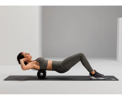 Woman in gray athletic wear lies on a yoga mat with a foam roller under her upper back, performing a thoracic spine stretch.