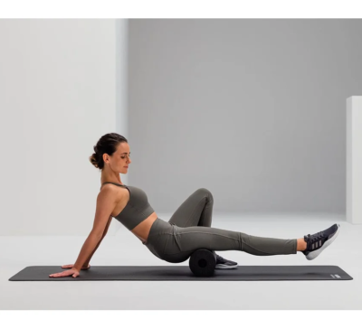 Woman in gray athletic wear uses a foam roller on a yoga mat, performing a back-/hip-mobility stretch in a bright studio.