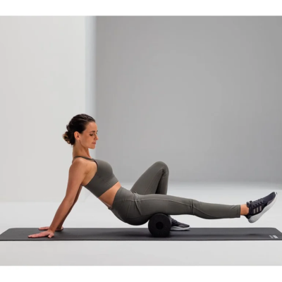 Woman in gray athletic wear uses a foam roller on a yoga mat, performing a back-/hip-mobility stretch in a bright studio.