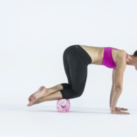 Woman in a pink sports bra and black leggings in aModified push-up position with a pink textured massage ball under her shins on a white studio backdrop.