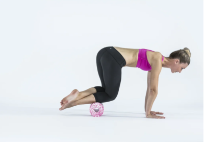 Woman in a pink sports bra and black leggings in aModified push-up position with a pink textured massage ball under her shins on a white studio backdrop.