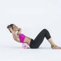 Woman in a pink sports bra and black leggings doing a crunch with a pink foam roller under her lower back on a white studio floor