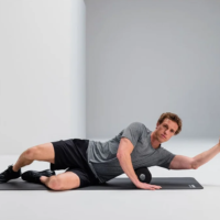Man lying on a black exercise mat, rolling on a foam roller with one arm extended forward for mobility exercise in a bright studio space.