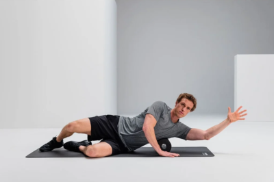 Man lying on a black exercise mat, rolling on a foam roller with one arm extended forward for mobility exercise in a bright studio space.